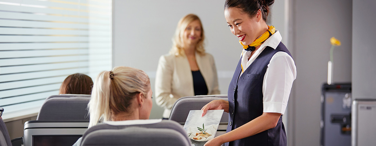 Eine Flugbegleiterin bietet in einer Trainingssituation einer Passagierin die Speisekarte an.