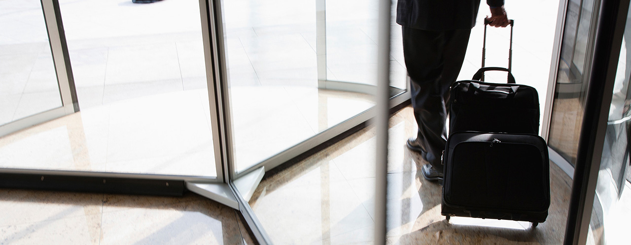 A pilot with a wheeled suitcase walks out through a revolving door