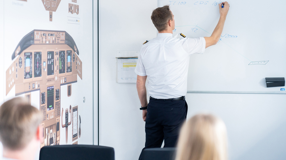 In einem Seminarraum schreibt ein Pilot als Ausbilder etwas an ein Whiteboard während zwei Personen zuschauen