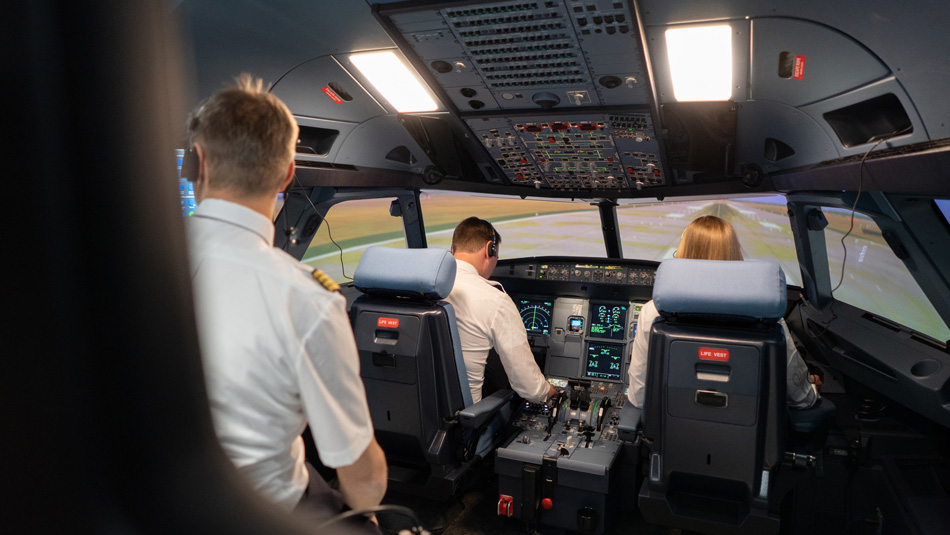 Two pilots sit in a cockpit simulator for training while a pilot trainer looks at a display