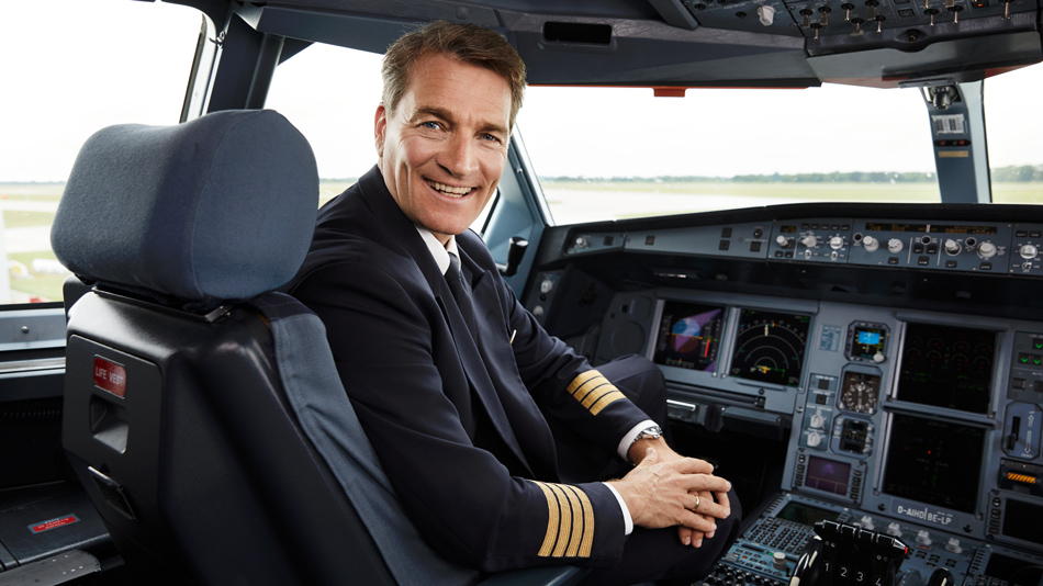 Ein Flugzeugkapitän sitzt in einem Cockpit und schaut lächelnd in die Kamera