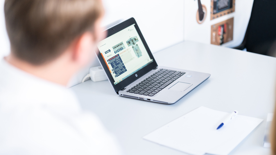 A notebook with a technical drawing on the screen stands on a table while two people look at it