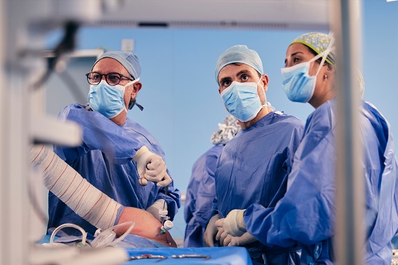 Mehrere Ärzt:innen in blauen Kitteln stehen an einem OP-Tisch und operieren