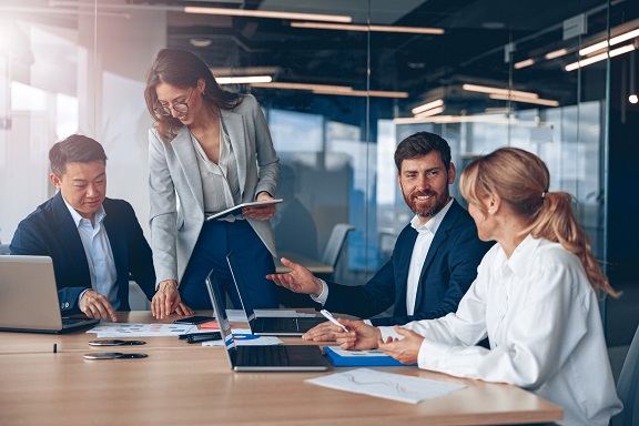 Vier Personen in Businesskleidung sitzen an einem Tisch mit Laptops und diskutieren.