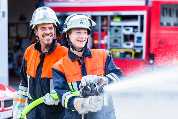 Zwei Feuerwehrleute mit Feuerwehruniform halten einen Schlauch in der Hand und löschen.