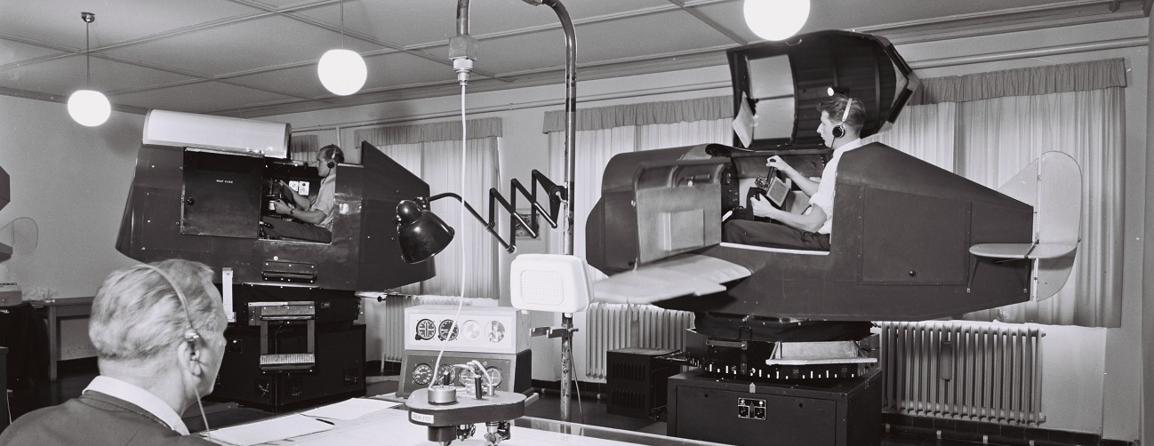 Black and white image showing two historical flight training devices, each with a pilot. In the foreground a trainer with a headset.