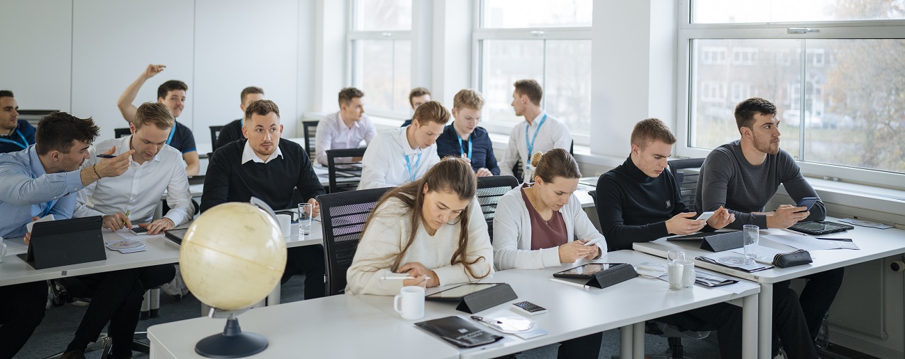 In a classroom, some individuals are sitting and working on tablets, while others are engaged in discussions.