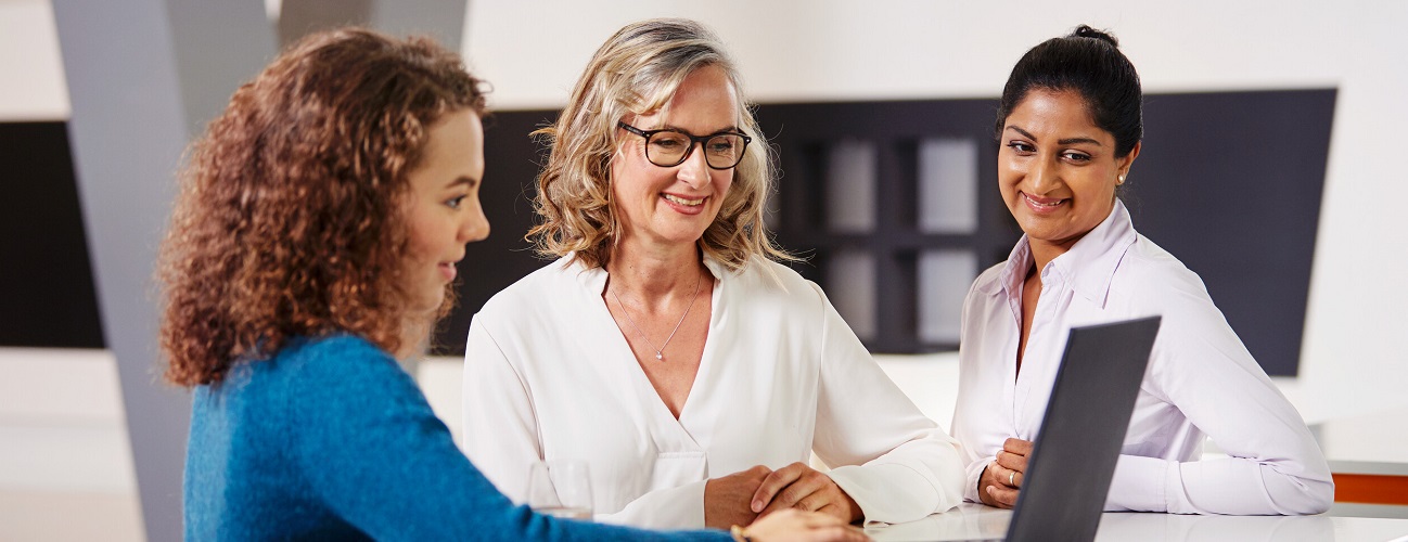  Three smiling people are standing around a table and looking at a laptop.