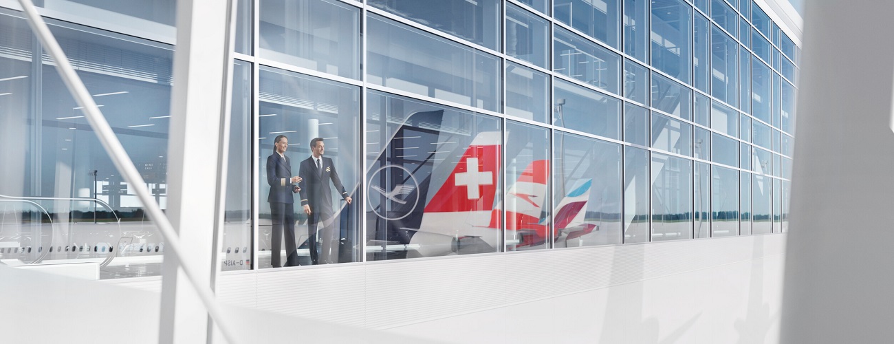 Two members of the cockpit crew stand behind a glass front. The aircraft tails with the liveries of Lufthansa, Swiss, Austrian and Eurowings are reflected in the windows.