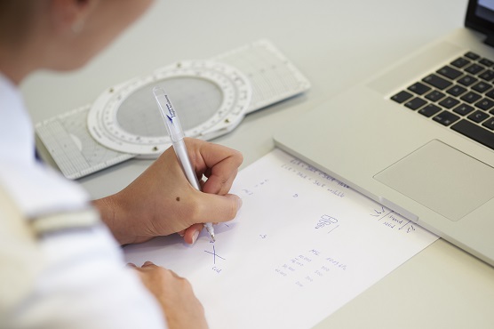 Eine Flugschüler:in sitzt vor einem geöffneten Laptop und macht Notizen auf einem Blatt Papier.