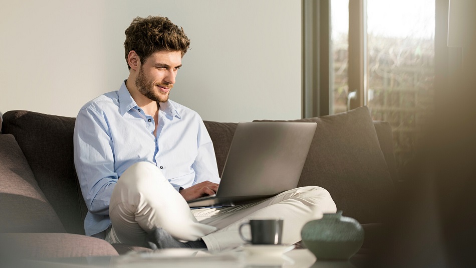 Eine Person sitzt auf einer Couch im Schneidersitz mit einem Laptop auf dem Schoss und tippt auf der Tastatur.