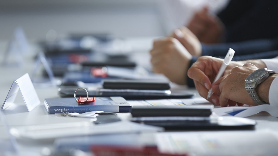  Several people sit at a table and write. Keyrings, calculators and notebooks are on the table.