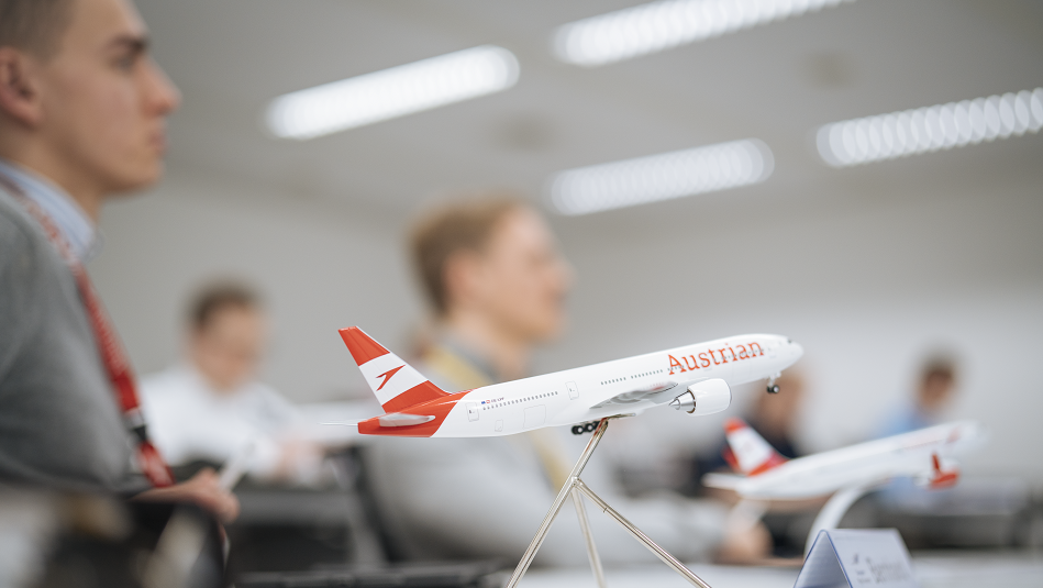 Im Vordergrund steht ein Modellflugzeug der Austrian, im Hintergrund sitzen Flugschüler:innen in einem Klassenzimmer