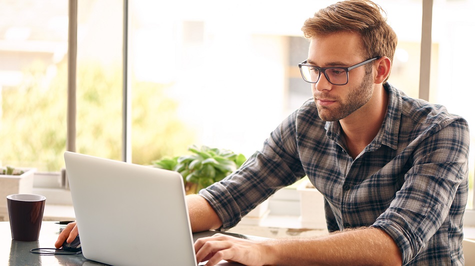 Eine Person sitzt an einem Tisch und arbeitet am Laptop, daneben steht ein Stapel Bücher.