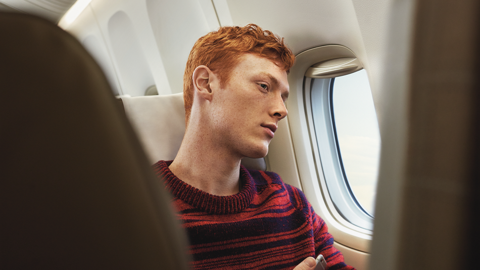 Eine Person mit roten Haaren sitzt im Flugzeug am Fenster, in einer Hand hält sie ein Handy.